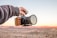 A hand is holding a professional camera with a wooden grip against a backdrop of a desert landscape at sunset. The sky is clear with subtle hues of orange and pink reflecting on the terrain.