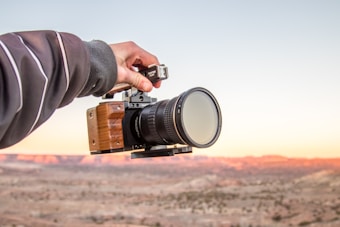 A hand is holding a professional camera with a wooden grip against a backdrop of a desert landscape at sunset. The sky is clear with subtle hues of orange and pink reflecting on the terrain.