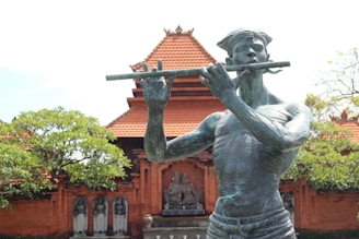 A bronze statue of a musician playing a flute is prominently displayed in front of a traditional temple-like structure with intricate red brickwork and a tiered roof. Surrounding the statue are lush green trees and additional stone carvings.