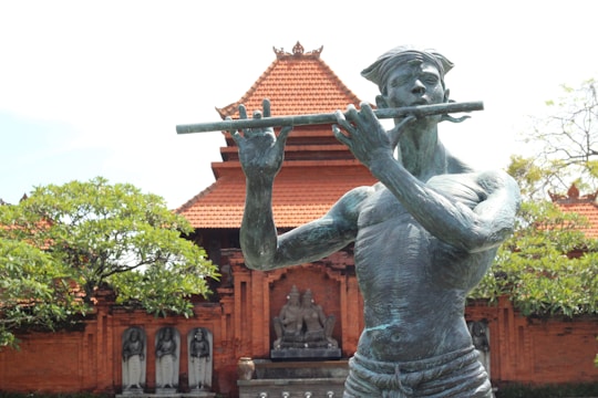 A bronze statue of a musician playing a flute is prominently displayed in front of a traditional temple-like structure with intricate red brickwork and a tiered roof. Surrounding the statue are lush green trees and additional stone carvings.