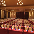 A large, well-lit conference room set up for an event with multiple rows of tables and chairs. Each table is covered with a maroon tablecloth and equipped with a notepad, pen, and a blue bottle of water. Elegant chandeliers and warm lighting create a cozy and professional atmosphere. The walls are decorated with soft, neutral tones enhancing the formal ambiance.