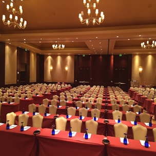 A large, well-lit conference room set up for an event with multiple rows of tables and chairs. Each table is covered with a maroon tablecloth and equipped with a notepad, pen, and a blue bottle of water. Elegant chandeliers and warm lighting create a cozy and professional atmosphere. The walls are decorated with soft, neutral tones enhancing the formal ambiance.