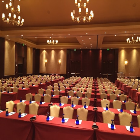 A large, well-lit conference room set up for an event with multiple rows of tables and chairs. Each table is covered with a maroon tablecloth and equipped with a notepad, pen, and a blue bottle of water. Elegant chandeliers and warm lighting create a cozy and professional atmosphere. The walls are decorated with soft, neutral tones enhancing the formal ambiance.
