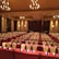 A large, well-lit conference room set up for an event with multiple rows of tables and chairs. Each table is covered with a maroon tablecloth and equipped with a notepad, pen, and a blue bottle of water. Elegant chandeliers and warm lighting create a cozy and professional atmosphere. The walls are decorated with soft, neutral tones enhancing the formal ambiance.
