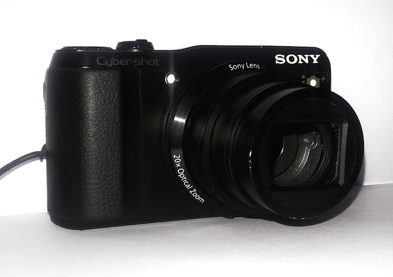A black digital camera with a textured grip and large lens is positioned on a plain surface. The brand name is clearly visible, along with a mention of '20x Optical Zoom' near the lens. The camera appears to be compact and designed for portability.
