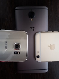 Three smartphones are placed on a dark wooden surface. The phones include models from OnePlus, Samsung, and Apple, each showing the rear side with branding visible. The arrangement highlights the camera modules and brand logos.