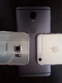 Three smartphones are placed on a dark wooden surface. The phones include models from OnePlus, Samsung, and Apple, each showing the rear side with branding visible. The arrangement highlights the camera modules and brand logos.