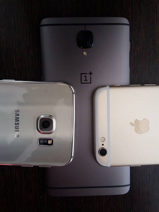 Three smartphones are placed on a dark wooden surface. The phones include models from OnePlus, Samsung, and Apple, each showing the rear side with branding visible. The arrangement highlights the camera modules and brand logos.