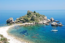 A small, lush island surrounded by clear blue water and rocky shores. A white sailboat is anchored near the island. The beach has a few people enjoying the scenery and sun. The vegetation on the island is dense and green.