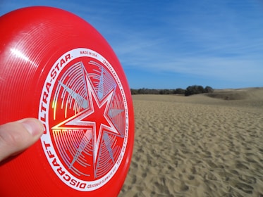 A red frisbee is held in a hand against a backdrop of sandy dunes under a clear blue sky. The frisbee features a stylized star design with the words 'Discraft Ultra-Star' printed on it.