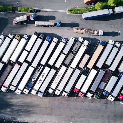 Aerial view of a parking lot filled with numerous trucks and trailers aligned in rows. The trucks are parked closely together, with notable variations in color and size. The ground is paved, and there are green areas and streets surrounding the parking space.