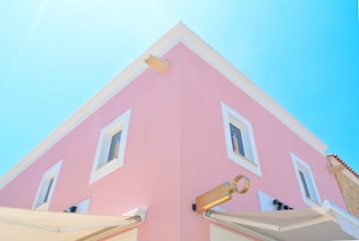 A two-story building with a pastel pink facade and white-framed windows. The architecture has a clean, modern style with a prominent corner and a sign resembling a key. The sky is a clear, bright blue, adding a sense of openness.