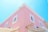 A two-story building with a pastel pink facade and white-framed windows. The architecture has a clean, modern style with a prominent corner and a sign resembling a key. The sky is a clear, bright blue, adding a sense of openness.