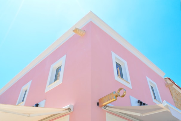 A two-story building with a pastel pink facade and white-framed windows. The architecture has a clean, modern style with a prominent corner and a sign resembling a key. The sky is a clear, bright blue, adding a sense of openness.