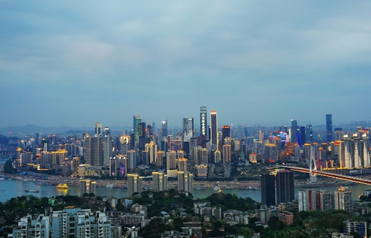 An expansive cityscape featuring a skyline filled with tall, illuminated skyscrapers. A river runs through the city, with a brightly lit bridge crossing it. The sky is overcast, casting a soft, diffused light across the scene. Buildings are densely packed, reflecting urban development.