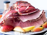 A platter featuring a large baked ham sliced into thin portions, arranged artistically. The ham has a glazed, caramelized outer layer, giving it a glossy appearance. Surrounding the meat are several slices of fresh apples, adding a touch of color to the presentation. In the background, a jar is partially visible, possibly containing spices.
