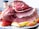 A platter featuring a large baked ham sliced into thin portions, arranged artistically. The ham has a glazed, caramelized outer layer, giving it a glossy appearance. Surrounding the meat are several slices of fresh apples, adding a touch of color to the presentation. In the background, a jar is partially visible, possibly containing spices.
