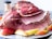 A platter featuring a large baked ham sliced into thin portions, arranged artistically. The ham has a glazed, caramelized outer layer, giving it a glossy appearance. Surrounding the meat are several slices of fresh apples, adding a touch of color to the presentation. In the background, a jar is partially visible, possibly containing spices.