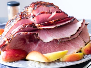 A platter featuring a large baked ham sliced into thin portions, arranged artistically. The ham has a glazed, caramelized outer layer, giving it a glossy appearance. Surrounding the meat are several slices of fresh apples, adding a touch of color to the presentation. In the background, a jar is partially visible, possibly containing spices.