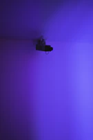 A surveillance camera is mounted on an indoor ceiling, illuminated by a purple and blue gradient light. The scene is minimalistic with a focus on the camera.