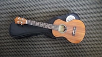 A wooden ukulele rests on a black soft case, placed on a patterned carpet. The ukulele features four strings, a light brown polished finish, and silver tuning pegs. A white sticker with a design is partially visible on the case.