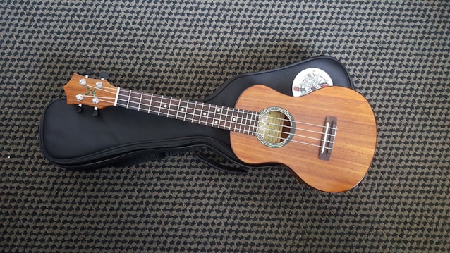 A wooden ukulele rests on a black soft case, placed on a patterned carpet. The ukulele features four strings, a light brown polished finish, and silver tuning pegs. A white sticker with a design is partially visible on the case.