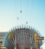 Three individuals are ziplining across the sky above a large, intricately designed metal structure with a curved canopy. The architecture appears modern with visible support beams and a mosaic-like pattern. In the foreground, a colorful sign with the words 'Trader Bill' is partially visible against a clear, pale blue sky.