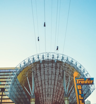Three individuals are ziplining across the sky above a large, intricately designed metal structure with a curved canopy. The architecture appears modern with visible support beams and a mosaic-like pattern. In the foreground, a colorful sign with the words 'Trader Bill' is partially visible against a clear, pale blue sky.