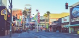 A bustling urban street scene featuring numerous neon signs and colorful advertisements. Prominent structures include a tall sign with the word 'Vegas' and another sign for a Coca-Cola store. Various storefronts and palm trees line the street, with a clear view down the road. Pedestrians can be seen walking, and the sky is clear.