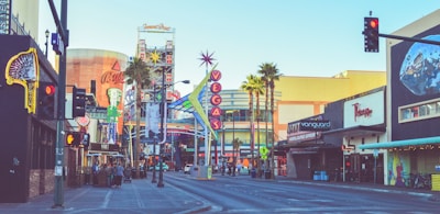 A bustling urban street scene featuring numerous neon signs and colorful advertisements. Prominent structures include a tall sign with the word 'Vegas' and another sign for a Coca-Cola store. Various storefronts and palm trees line the street, with a clear view down the road. Pedestrians can be seen walking, and the sky is clear.
