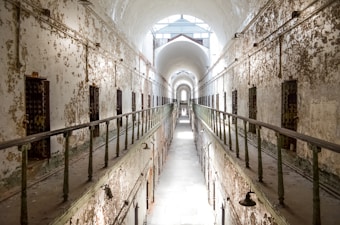 A long, narrow corridor with peeling paint, high arched ceilings, and prison cell doors lined along the walls. Metal railings run down both sides of the walkway, and natural light pours in from a skylight, casting shadows.