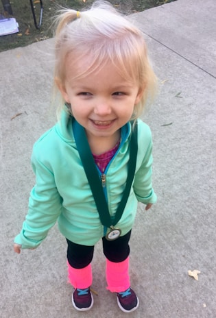 A young child with blond hair stands on a concrete surface, wearing a light green jacket and a medal around their neck. The child is smiling and has bright pink leggings over black pants and dark sneakers.
