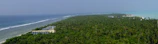 A vast expanse of lush, green palm trees stretches out towards the horizon, bordered by a deep blue ocean on one side and a light turquoise sea on the other. To the left, there are visible solar panels among the vegetation. In the distance, there are a few buildings near the shoreline.