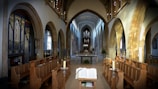 An ornate church interior with tall arches and a vaulted ceiling. Wooden pews line the aisle leading to an altar positioned in front of a large pipe organ. The lighting is soft, streaming through stained glass windows, illuminating the space warmly. A Bible is open on a stand in the foreground.