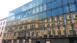 A modern glass building with an intricate facade features a reflection of a classic architectural structure with arched windows and beige-colored walls. The glass panels create a multifaceted effect, blending contemporary and traditional architectural styles.