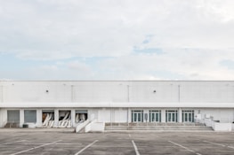 A large industrial warehouse with white exterior walls and multiple garage doors. The building is devoid of any signage or decoration, and several air conditioning ducts extend from some of the open bays. The surrounding area is an empty parking lot with marked spaces, and the sky is overcast with a few patches of blue.