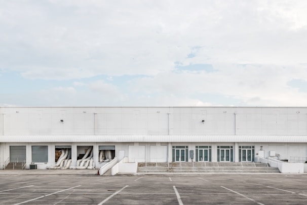 A large industrial warehouse with white exterior walls and multiple garage doors. The building is devoid of any signage or decoration, and several air conditioning ducts extend from some of the open bays. The surrounding area is an empty parking lot with marked spaces, and the sky is overcast with a few patches of blue.