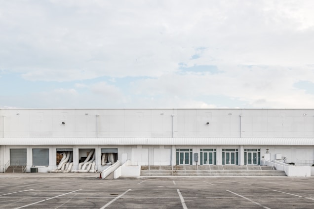 A large industrial warehouse with white exterior walls and multiple garage doors. The building is devoid of any signage or decoration, and several air conditioning ducts extend from some of the open bays. The surrounding area is an empty parking lot with marked spaces, and the sky is overcast with a few patches of blue.