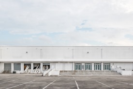 A large industrial warehouse with white exterior walls and multiple garage doors. The building is devoid of any signage or decoration, and several air conditioning ducts extend from some of the open bays. The surrounding area is an empty parking lot with marked spaces, and the sky is overcast with a few patches of blue.