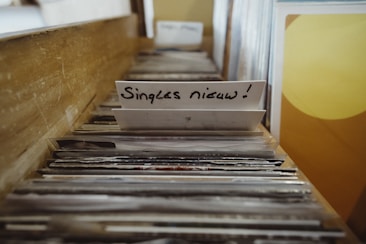 A collection of vinyl records stored in a wooden crate, with a focus on a divider labeled 'Singles nieuw!' indicating new singles. The records are arranged vertically, with album covers visible at the top. The scene gives a sense of browsing through a record collection.