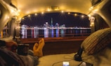 A cozy scene inside a van with a view of a city skyline at night. String lights illuminate the interior, creating a warm and inviting atmosphere. A person relaxes with their feet stretched out towards the city view, holding a cup. A pillow and headphones lie nearby, with a smartphone displaying content.