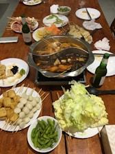 A hot pot meal set up on a wooden table, featuring a divided pot with broth and various ingredients such as tofu, quail eggs, and vegetables on skewers. Surrounding the pot are plates filled with an assortment of raw ingredients including leafy greens, fish balls, tofu cubes, sliced meats, and edamame. Condiments, bottles of beer, plates, and napkins are also visible.