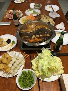 A hot pot meal set up on a wooden table, featuring a divided pot with broth and various ingredients such as tofu, quail eggs, and vegetables on skewers. Surrounding the pot are plates filled with an assortment of raw ingredients including leafy greens, fish balls, tofu cubes, sliced meats, and edamame. Condiments, bottles of beer, plates, and napkins are also visible.