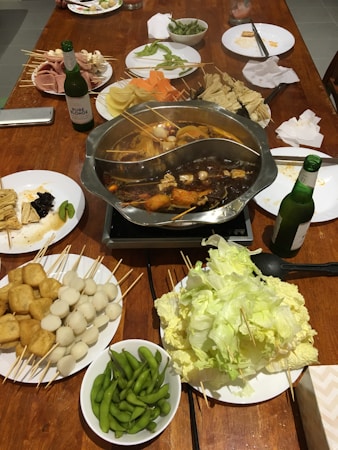 A hot pot meal set up on a wooden table, featuring a divided pot with broth and various ingredients such as tofu, quail eggs, and vegetables on skewers. Surrounding the pot are plates filled with an assortment of raw ingredients including leafy greens, fish balls, tofu cubes, sliced meats, and edamame. Condiments, bottles of beer, plates, and napkins are also visible.