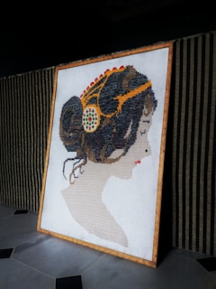 A framed cross-stitch artwork depicting a profile of a woman with intricately styled hair and decorative elements. The hairstyle includes colorful accents, and the background is a woven pattern with vertical stripes.