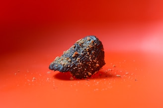 A dark chocolate-covered treat is sprinkled with crushed nuts, set against a vibrant orange background. Crumbs are scattered around the base.