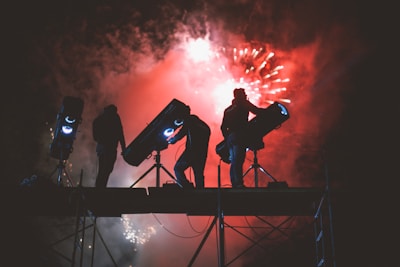 Technicians coordinating pyrotechnic effects with flames and drones illuminating the night sky.