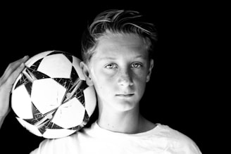 A black and white portrait of a young person holding a soccer ball against a dark background. The expression is neutral, and the focus is sharp on both the face and the ball.
