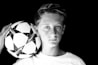 A black and white portrait of a young person holding a soccer ball against a dark background. The expression is neutral, and the focus is sharp on both the face and the ball.