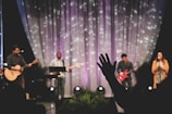 A musical performance on stage features a group of musicians, each with an instrument. The backdrop includes softly lit drapery with scattered light patterns. A person in the foreground raises a hand in what seems to be an expression of involvement or enjoyment.